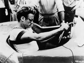 James Dean sits behind the wheel of a sports car in a still from ‘The James Dean Story,’ 1957. (Credit: Warner Bros./Getty Images)