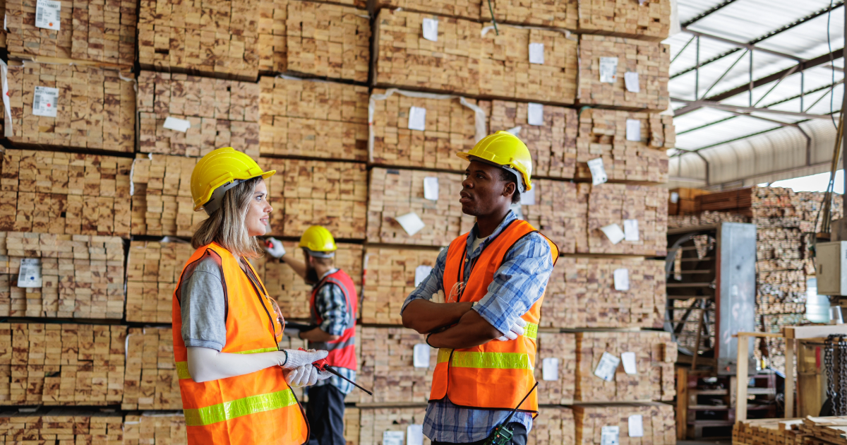 Employees standing inside a building materials retail location surrounded by stacked lumber inventory.