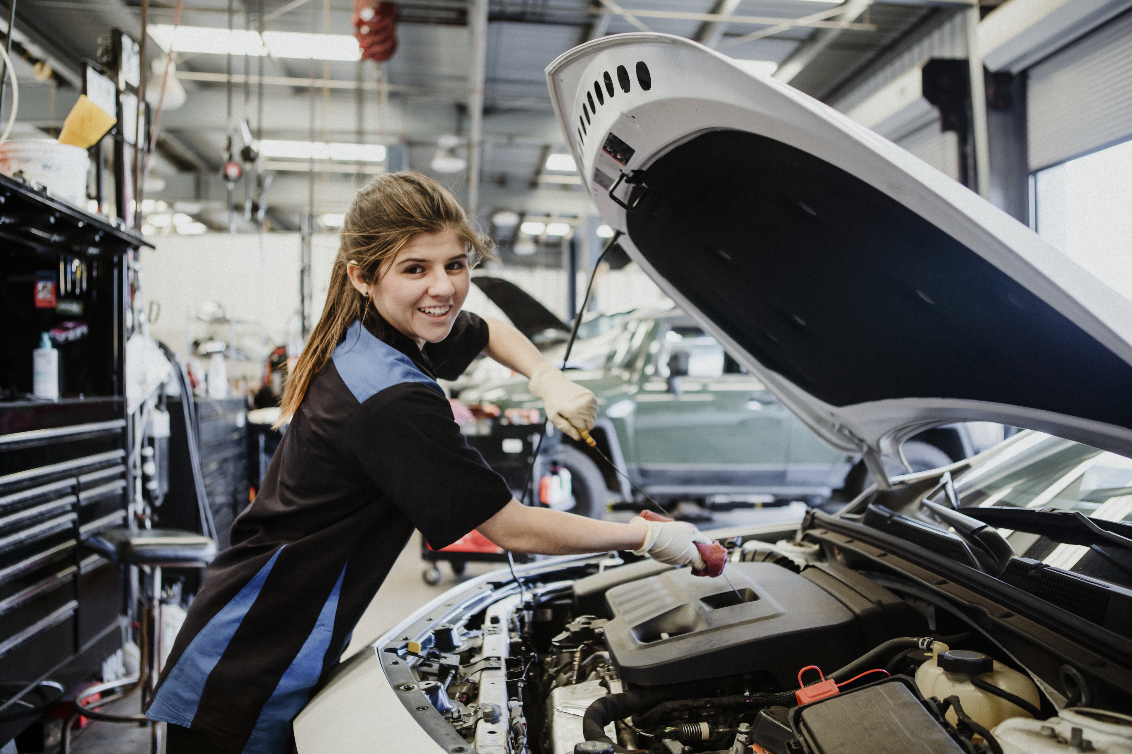 Young female mechanic smiling at camera while working under a car's hood inside an auto service center.
